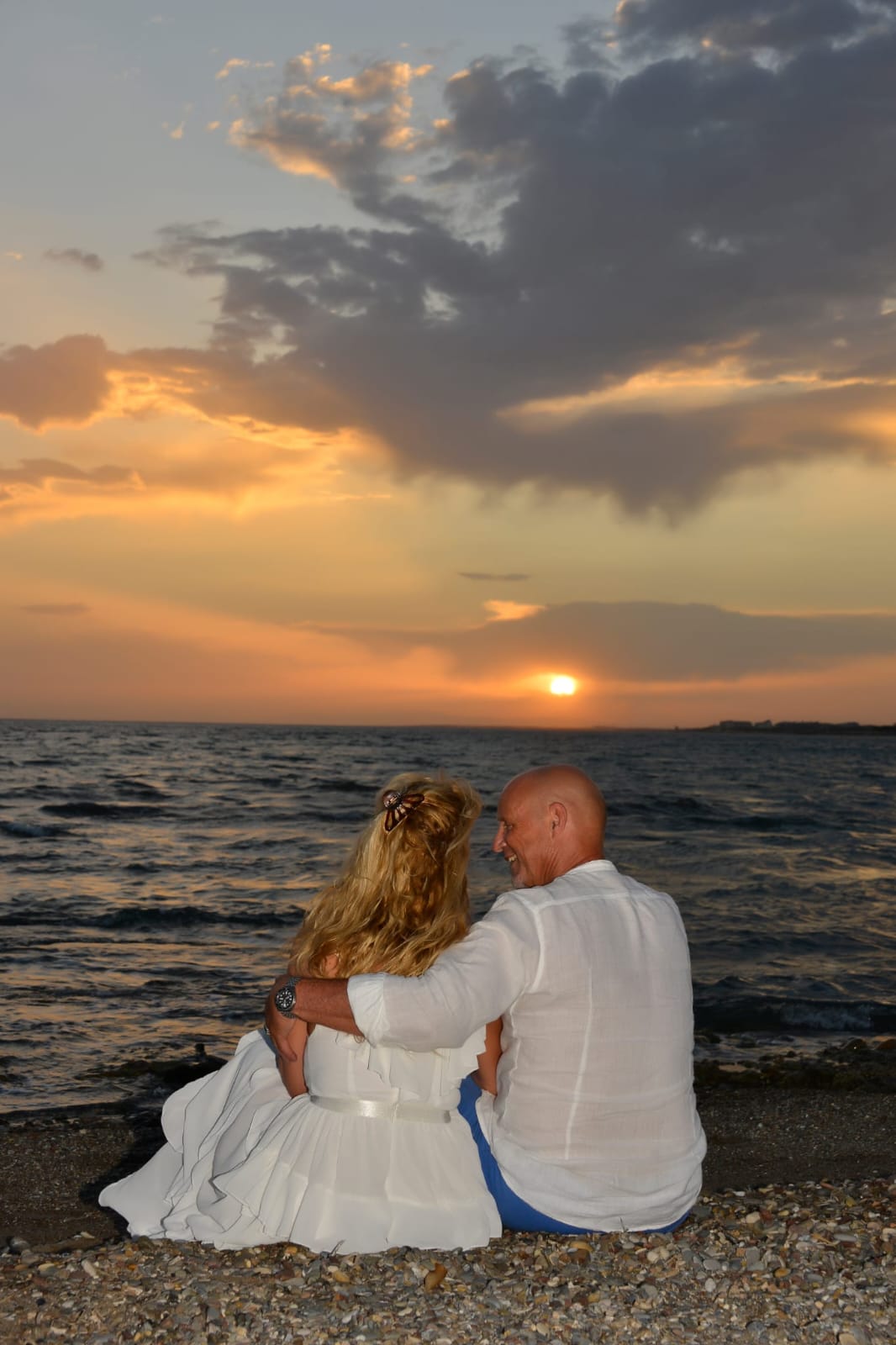 Pascale, de coach, en haar man Remy zitten naast elkaar bij zonsondergang aan de zee